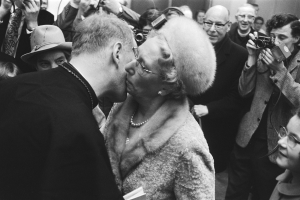 Ein Mann und eine Frau umarmen und küssen sich vor einer Menge, einige halten Kameras, dargestellt in Schwarz-Weiß.