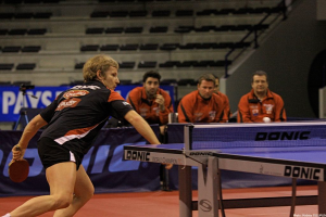 Ein Mann hält einen Tischtennisschläger und steht vor einem Tischtennistisch mit vier im Hintergrund sitzenden Personen.