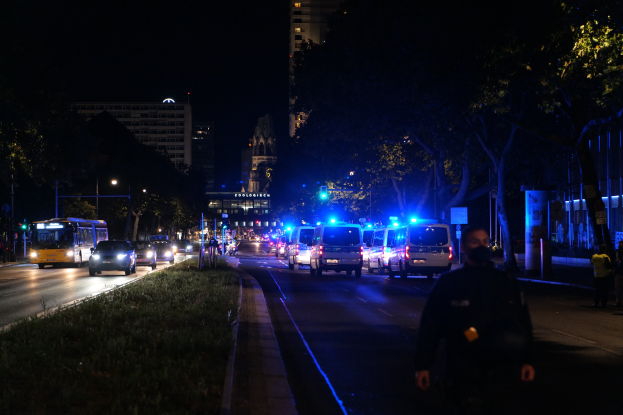 Eine nächtliche Straßenansicht mit Polizeiwagen und Bussen, die fahren, Menschen auf dem Bürgersteig gehen, Gras auf der linken Seite, Bäume und Gebäude im Hintergrund und Laternen, die die Straße beleuchten.
