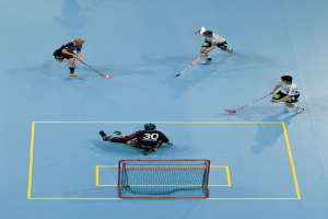 Drei Personen in Sportbekleidung spielen Hockey, wobei eine Person auf der rechten Seite den Ball mit einem Hockeystock schlägt und eine Person hinter einem Netz auf dem Boden sitzt.