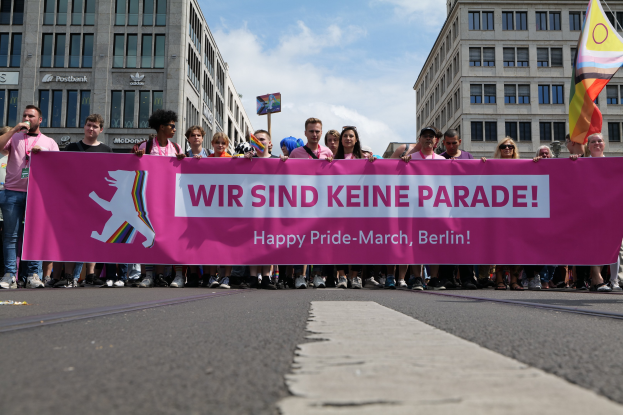 Eine Gruppe von Menschen bei einer Pride-Veranstaltung in Berlin, die eine pinkfarbene Fahne mit der Aufschrift "Happy Pride March Berlin" halten, einige tragen Mützen und Schutzbrillen, einer hält eine Fahne, vor einem Hintergrund von Gebäuden, einem Schild und einem bewölkten Himmel.