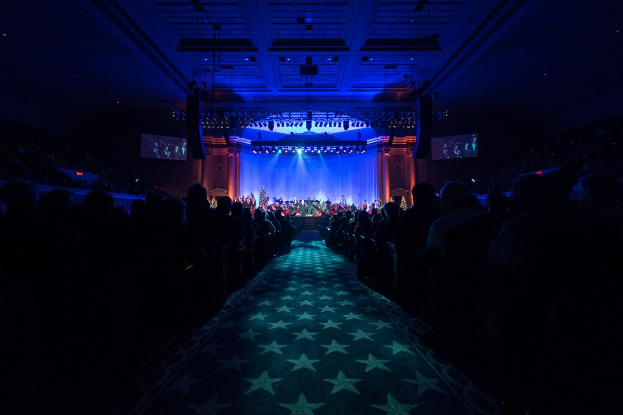 Ein großer, dämmrig beleuchteter Auditorium mit Menschen in Reihen vor einer Bühne, auf der Musiker spielen, mit Deckenleuchten und Fernsehgeräten, was auf einen Konzertsaal während der 2019 National Convention hinweist.