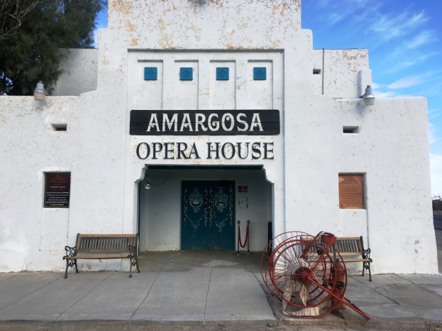 Das Amargosa Opernhaus in San Diego, Kalifornien, ein weißes Gebäude mit einer grünen Tür und zwei Bänken davor, flankiert von zwei Schildern mit Schrift, mit einem roten Objekt auf dem Gehweg und einem Baum links, unter einem bewölkten Himmel.