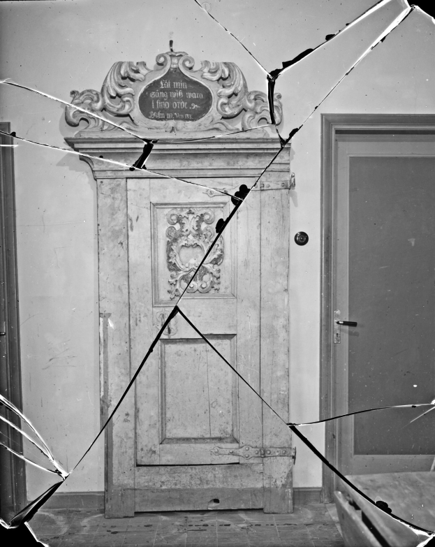 Schwarzes und weißes Foto einer kaputten Tür in einem Raum, mit zerbrochenem Glas drumherum und einer Wand im Hintergrund.