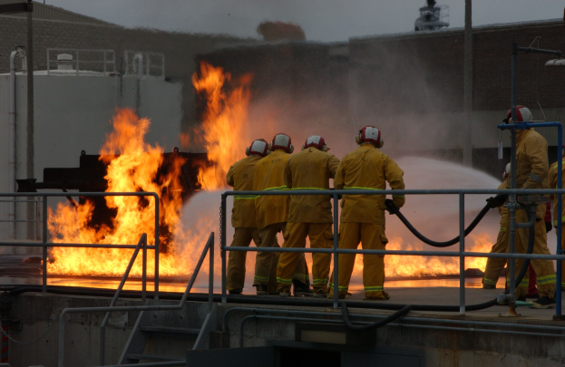 Feuerwehrleute in Helmen auf einem Dach mit Schläuchen stehend, mit Geländern und Treppen darunter und einem Gebäude und Himmel im Hintergrund.