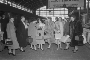 Schwarzes und weißes Bild einer Gruppe von Frauen, die an einem Bahnhof stehen und Taschen halten, mit einem Zug auf der linken Seite und Geländern und einer Tafel auf der rechten Seite.