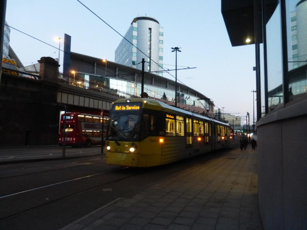 Ein Bus fährt auf einer Straße mit Gebäuden auf beiden Seiten, jedes mit Glasfenstern, und einer Brücke auf der linken Seite. Es gibt auch Laternenmasten mit Lampen entlang der Straße.