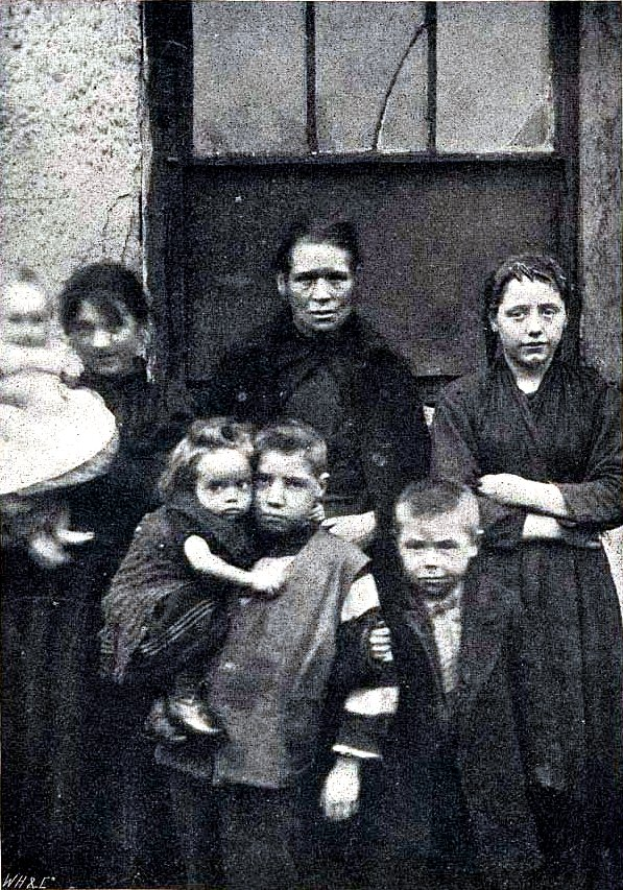 Ein altes Schwarz-Weiß-Foto einer Familie, die vor einer Wand mit einem Fenster posiert, mit dem Text "Mutter Teresa mit ihren Kindern" unten.