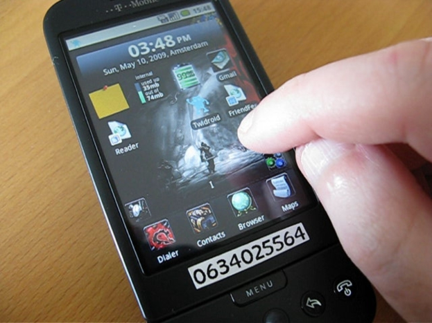 Eine Person nutzt ein Mobiltelefon auf einem Tisch, mit mehreren Apps auf dem Bildschirm und einem Zettel mit einer Nummer am Telefon.