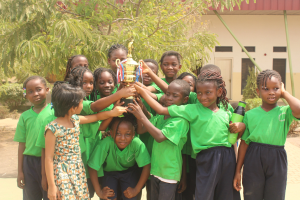 Gruppe von Kindern mit einem Pokal auf einem Schulhof mit Bäumen und einem Gebäude im Hintergrund.