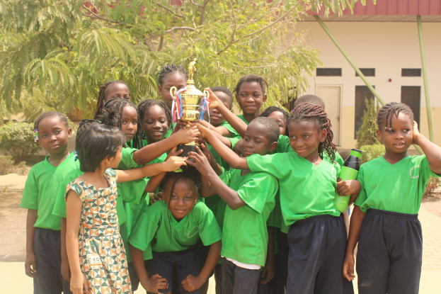 Gruppe von Kindern mit einem Pokal auf einem Schulhof mit Bäumen und einem Gebäude im Hintergrund.