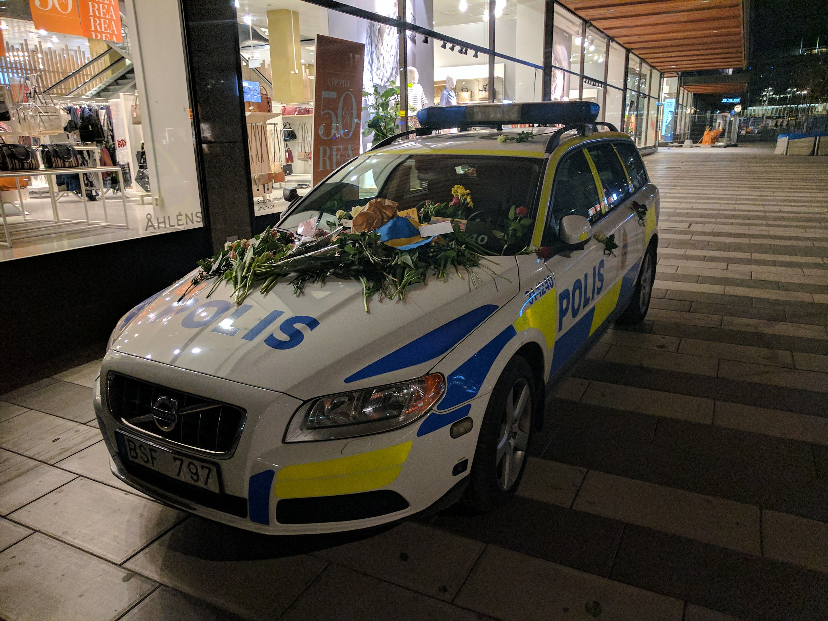 Ein mit Blumen und Blättern geschmückter Polizeiwagen parkt nachts vor einem Geschäft mit Glaswänden, das Puppen in Kleidern und beleuchtete Schilder zeigt, mit Bäumen und Gebäuden im Hintergrund.