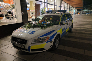 Ein mit Blumen und Blättern geschmückter Polizeiwagen parkt nachts vor einem Geschäft mit Glaswänden, das Puppen in Kleidern und beleuchtete Schilder zeigt, mit Bäumen und Gebäuden im Hintergrund.