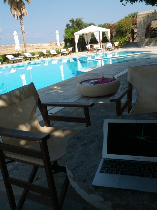 Ein Schwimmbadbereich mit Stühlen, einem Tisch mit einem Laptop, einer Topfpflanze mit Blütenblättern, Palmen, anderen Bäumen, einem Unterstand und einem Gebäude im Hintergrund unter einem klaren Himmel.