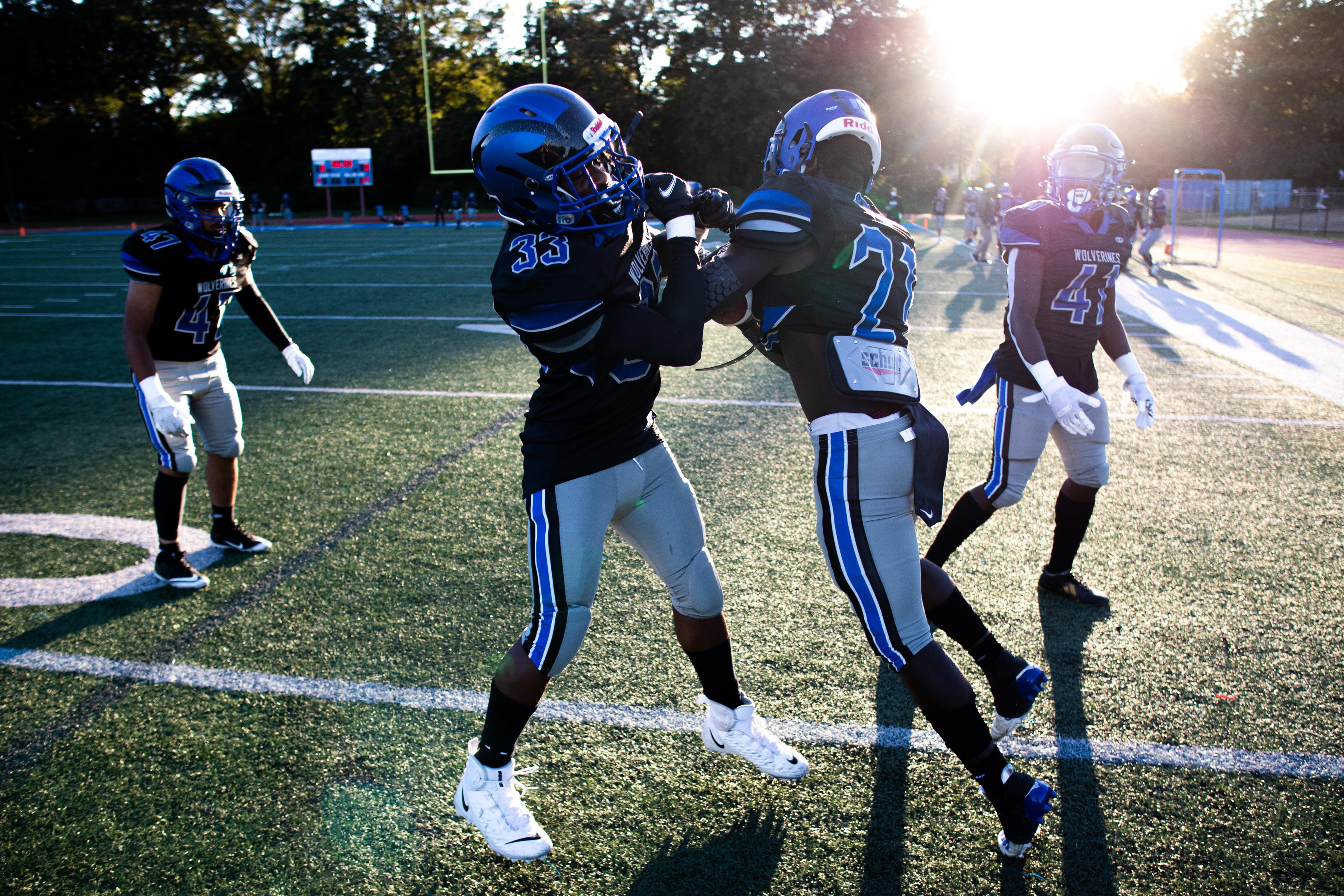 Junge Männer spielen Fußball auf einem Feld mit Bäumen und einem klaren Himmel, tragen Helme und Stollenschuhe.