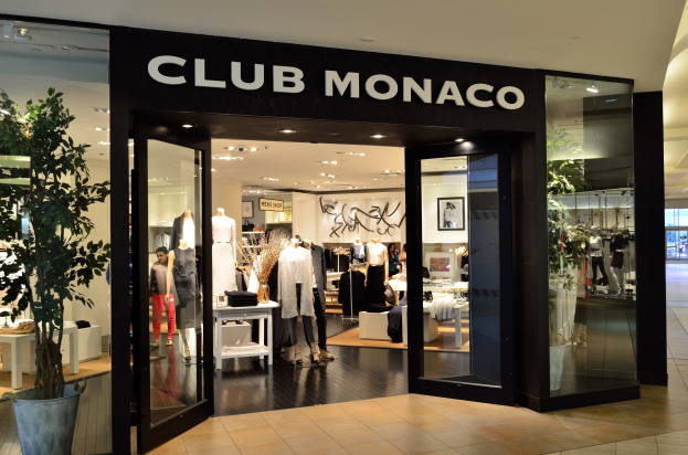 Vorderfront eines Club Monaco Einzelhandelsgeschäfts in einem Einkaufszentrum, mit Glaswänden und -türen, Schaufensterpuppen in Kleidern, Auslagetischen mit Waren, einem Topf mit einer Pflanze und Deckenbeleuchtung.