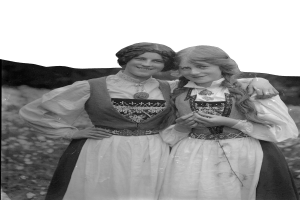 Zwei junge Frauen in traditioneller bayrischer Tracht, mit weißen und schwarzen Kleidern mit aufwendiger Stickerei und Perlen, posieren vor der Kamera und lächeln in die Kamera. Der Hintergrund besteht aus üppigen grünen Pflanzen und einem hellen weißen Himmel.