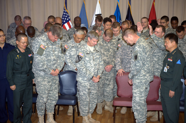 Eine Gruppe von Menschen, mostly in military dress, stehen zwischen roten und blauen Stühlen mit Flaggen im Hintergrund.