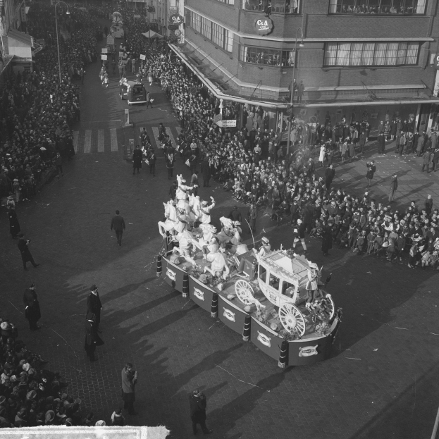 Eine große Menschenmenge umringt einen Festwagen bei einem Umzug, der mit bunten Luftballons und Bändern geschmückt ist, mit Gebäuden und Laternenmasten im Hintergrund, in Schwarz-Weiß.