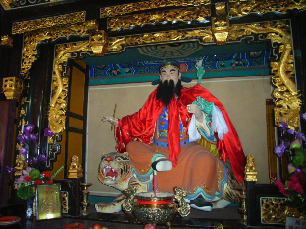 Eine Statue eines chinesischen Mönchs in bunter Kleidung sitzt auf einer Tigerstatue innerhalb eines Bogens mit goldenen Verzierungen, vorne ein Blumenstrauß und in der Mitte eine Schale.