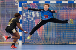 Zwei Frauen beim Handballspielen auf einem Platz mit einem Tor und einer Fahne im Hintergrund.