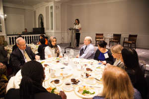 Eine Gruppe von Menschen sitzt um einen Esstisch mit Tellern, Bestecken und Trinkgefäßen, während eine Frau in der Nähe eines Podiums mit einem Mikrofon im Hintergrund steht.