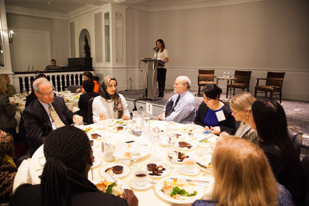 Eine Gruppe von Menschen sitzt um einen Esstisch mit Tellern, Bestecken und Trinkgefäßen, während eine Frau in der Nähe eines Podiums mit einem Mikrofon im Hintergrund steht.