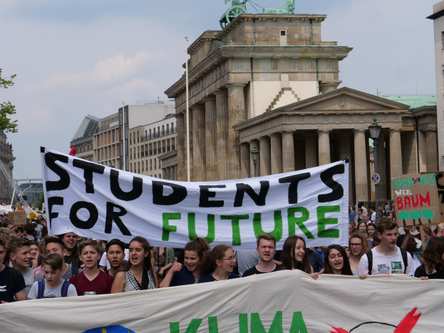 Gruppe von Schülern marschiert in Berlin mit einer bunten "Students for Future"-Schlagzeile an Gebäuden, Bäumen und Himmel vorbei.