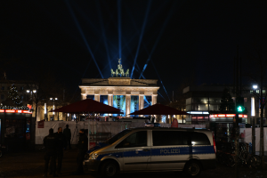 Ein Polizeiwagen steht nachts vor dem Reichstagsgebäude in Berlin, umgeben von ein paar Menschen und verschiedenen Gegenständen wie Schirmen, Gebäuden, Bäumen, Lichtern, Pfosten und einer Ampel.