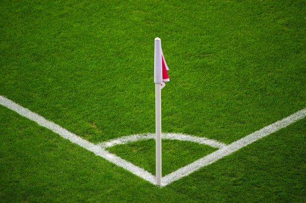 Ein Fussballfeld mit grüner Rasen, weissen Begrenzungslinien und einem Elfmeterpunkt, der mit einem weissen Pfahl mit einer rot-weissen Fahne markiert ist.
