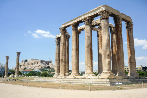 Alter Tempel von Olympian Zeus in Athen, Griechenland, mit seinen verbliebenen Säulen, umgeben von Bäumen und Felsen, vor einem bewölkten Himmel mit einer Festung im Hintergrund.