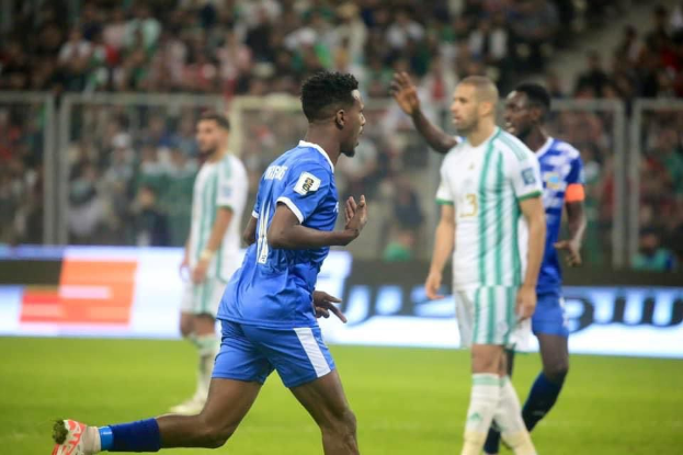 Ein Fussballspieler in einem blauen und weissen Trikot feiert mit erhobenen Armen, umgeben von einer Gruppe in weissen und grünen Trikots, mit einem unscharfen Hintergrund, der ein weisses und blaues Board und Geländer zeigt.