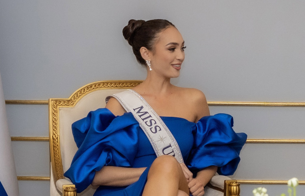 Frau in einem blauen Kleid mit einer "Miss Universe 2019" Schärpe, die auf einem Stuhl sitzt und von Blumen umgeben ist und lächelt.