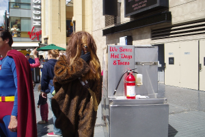 Eine Gruppe von Menschen, einige in Kostümen, versammelte sich vor einem Gebäude mit sichtbarer Beschriftung und Türen, mit einer Box und einem Feuerlöscher in der Nähe.