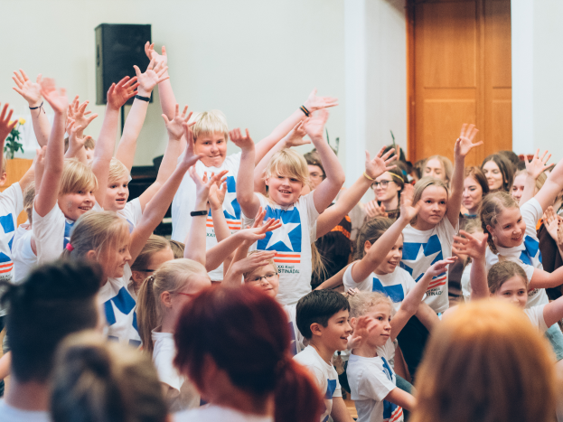 Eine Gruppe von Kindern steht mit erhobenen Händen vor einer Menge, mit einer Wand und einer Tür im Hintergrund; ein Tisch mit einer Blumenvase und einem Lautsprecher ist links zu sehen, was auf ein Sommerlager in Schweden hindeutet.