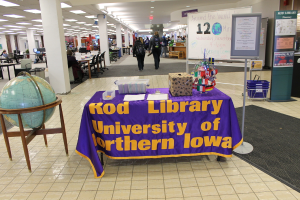 Eine Ausstellung mit einem Tisch, auf dem verschiedene Landesflaggen, eine Box, Papiere und ein Banner stehen, eine Kugel links daneben, ein Anschlagbrett rechts daneben mit ein paar Menschen daneben und mehreren Tischen und Stühlen links.
