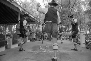 Eine Gruppe von Männern in traditioneller bayrischer Tracht tanzt auf einer Bühne vor einem gemischten Publikum, mit Bäumen und einer Hütte im Hintergrund, dargestellt in Schwarz-Weiß.