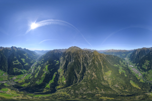 Panoramischer Blick auf ein Gebirgsmassiv von einem Hügel aus, mit grünem Gras, einer gewundenen Straße und einem hellblauen Himmel unter einer strahlenden Sonne.