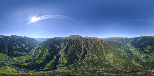 Panoramischer Blick auf ein Gebirgsmassiv von einem Hügel aus, mit grünem Gras, einer gewundenen Straße und einem hellblauen Himmel unter einer strahlenden Sonne.