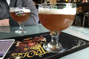 Zwei Gläser goldfarbenes Bier auf einem Tisch neben einem Handy, mit Menschen im Hintergrund und gerahmten Kunstwerken an der Wand.