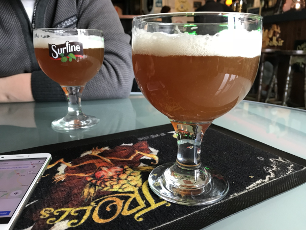 Zwei Gläser goldfarbenes Bier auf einem Tisch neben einem Handy, mit Menschen im Hintergrund und gerahmten Kunstwerken an der Wand.