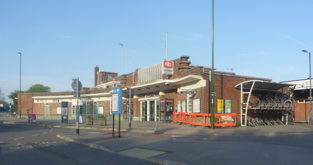 Ein belebter Bahnhof mit zahlreichen Fahrrädern davor, umgeben von Gebäuden, Straßeninfrastruktur, Kraftfahrzeugen, Bäumen und einem bewölkten Himmel.
