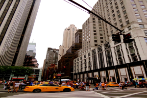 Eine Stadtstraße mit Gebäuden, Ampeln, Fenstern, wenigen Passanten, Autos und einem Himmel darüber.