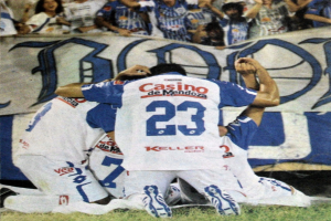 Eine Gruppe von Füßtballspielern feiert ein Tor auf dem Feld mit jubelnden Fans und einem Banner im Hintergrund.