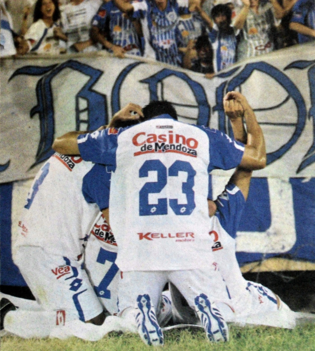 Eine Gruppe von Füßtballspielern feiert ein Tor auf dem Feld mit jubelnden Fans und einem Banner im Hintergrund.