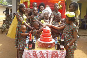 Eine Gruppe von Menschen steht um einen Tisch mit einem Kuchen, Flaschen und anderen Gegenständen in einer ländlichen Umgebung mit Häusern, Bäumen und einem klaren blauen Himmel, um einen Geburtstag zu feiern, wobei einige Hände halten und andere lächeln.