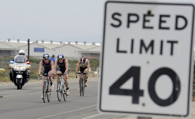 Eine Gruppe von Radfahrern mit Helmen fährt auf einer Straße neben einem Geschwindigkeitsbegrenzungsschild, mit Gebäuden, Masten, Pflanzen, Bäumen und einem klaren blauen Himmel im Hintergrund.