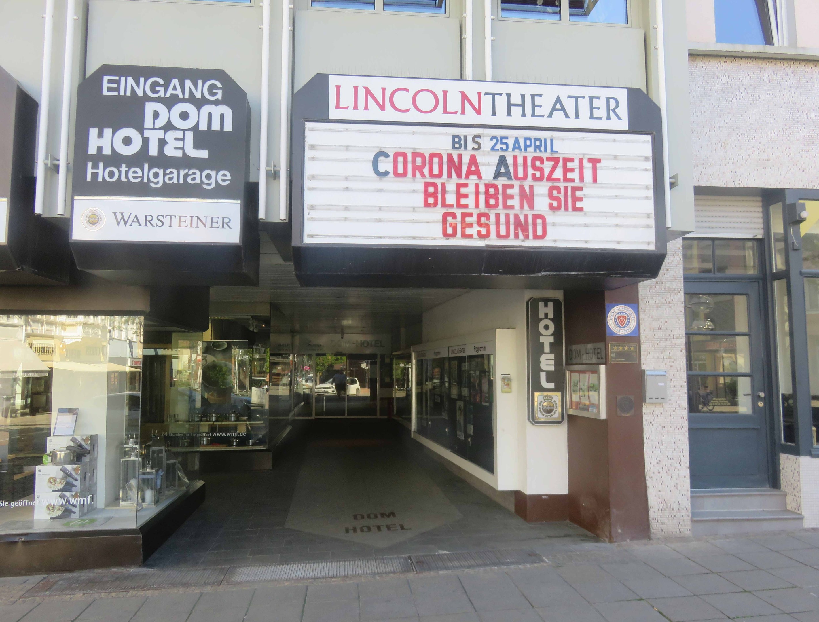 Außenansicht des Lincoln Theaters in Berlin, Deutschland, mit Glasfenstern und -türen sowie einer Tafel mit Text und einem Inneren, das eine pulsierende Stadtlandschaft vermuten lässt.