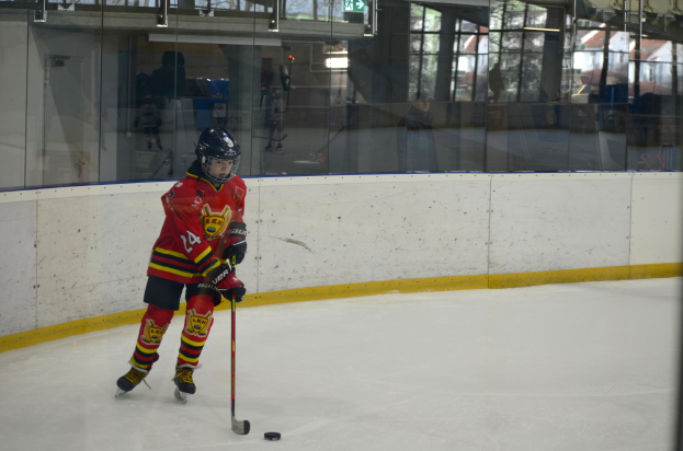 Ein junger Junge spielt Hockey auf einem Eisplatz, trägt einen Helm und hält einen Hockeystock, mit Menschen, Bäumen, Gebäuden und dem Himmel im Hintergrund.