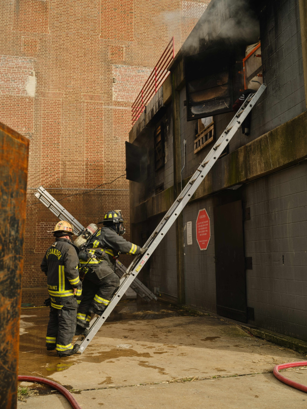 Feuerwehrleute in Helmen, die eine Leiter zu einem raucherfüllten Gebäude hinaufklettern, mit Metallobjekten links und Rohren an der Basis.